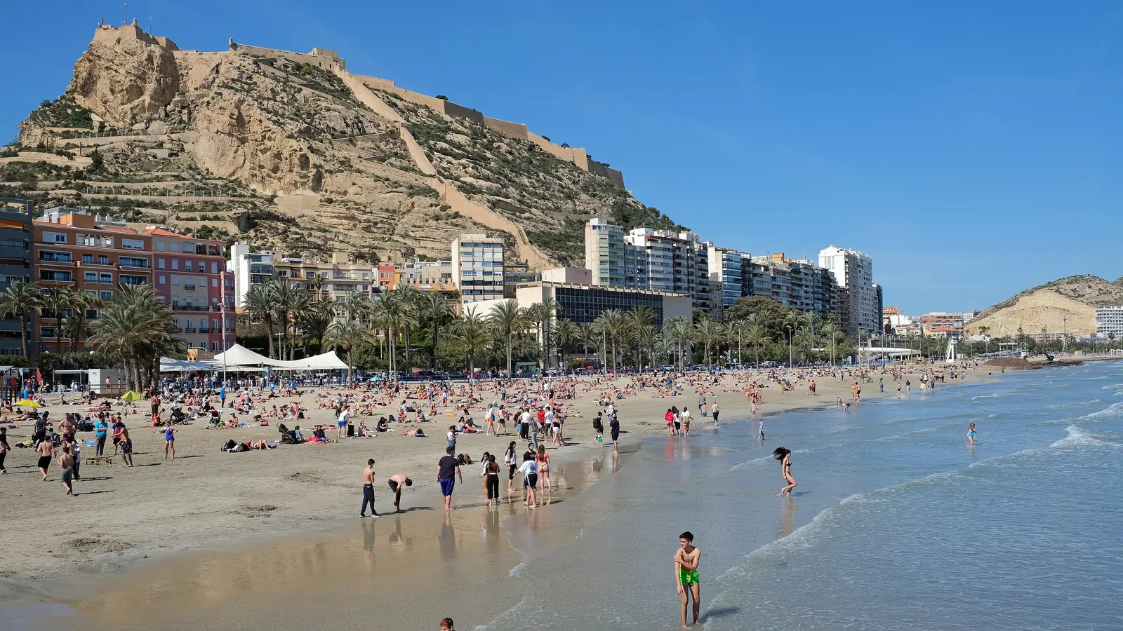 Santa Bárbara Castle on its rocky hill above Postiguet beach in Alicante, with swimmers at the edge of the sea and apartment blocks along the shore