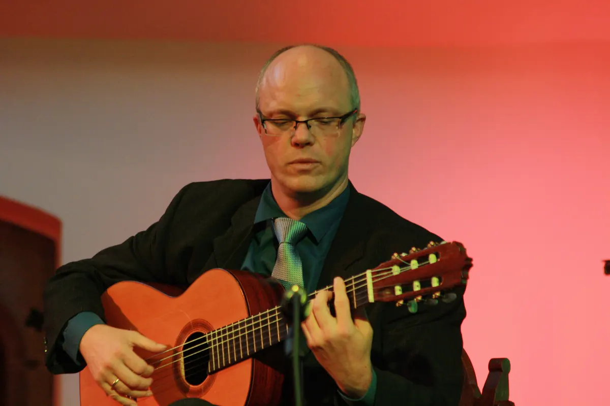 A guitarist in a dark jacket and green shirt plays a flamenco guitar on a stage with a red backdrop