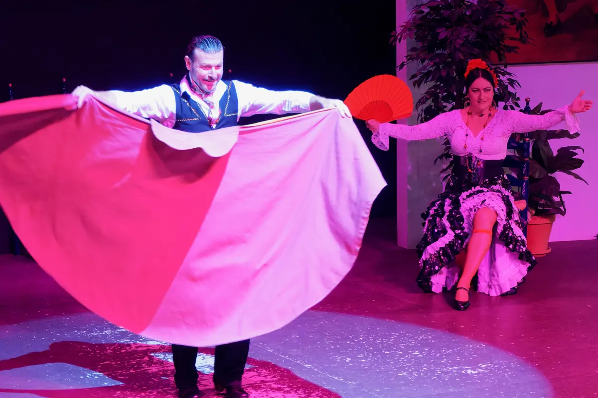 A male dancer spreads a large red and white cape wide on a spotlit stage while a female dancer in a black polka-dot dress sits with a red fan raised